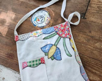 May include: A white tote bag with a patchwork floral design and a small pocket. The bag has a long white strap and a circular patch with the text "Moon Gold Goods". A woven basket sits in the background.