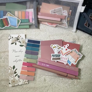 May include: A set of mesh pouches filled with colorful sticky notes, pens, and stickers. The pouches are labeled with the text "New day new chapter" and "Just one more chapter".