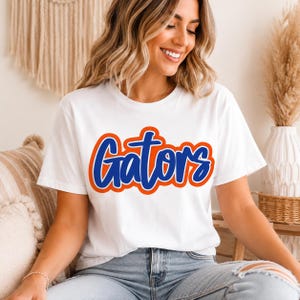 May include: A white short-sleeved t-shirt featuring the word Gators printed in a bold blue script font with a thick orange outline. The design is centered on the chest of the garment, which is paired with light blue denim jeans.