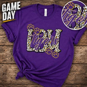 May include: Purple t-shirt with the text "Game Day" in white and yellow. The shirt features the letters "BDH" and the word "Tigers" in a leopard print design, with yellow paw prints.