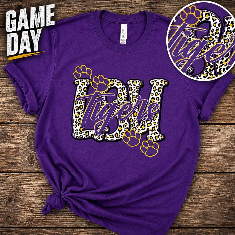 May include: Purple t-shirt with the text "Game Day" in white and yellow. The shirt features the letters "BDH" and the word "Tigers" in a leopard print design, with yellow paw prints.