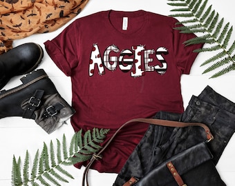 May include: A maroon t-shirt with the word "Aggies" in white letters with a red and black plaid and white polka dot pattern.