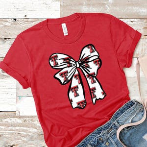 May include: Red t-shirt featuring a large white bow with a repeating pattern of the letter "T" in red and black. The shirt has short sleeves and a crew neck. The shirt is paired with denim shorts and a pink purse.
