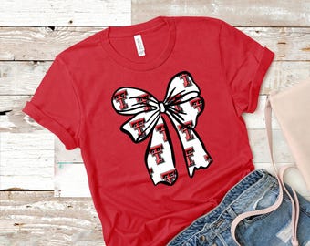May include: Red t-shirt featuring a large white bow with a repeating pattern of the letter "T" in red and black. The shirt has short sleeves and a crew neck. The shirt is paired with denim shorts and a pink purse.