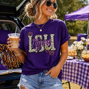 May include: A purple t-shirt with the text "LSU Tigers" in a leopard print design. The shirt has rolled-up sleeves and is paired with denim shorts. The person is wearing sunglasses and holding a beverage.