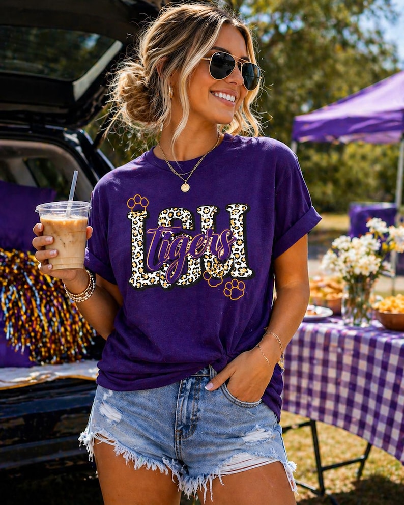 May include: A purple t-shirt with the text "LSU Tigers" in a leopard print design. The shirt has rolled-up sleeves and is paired with denim shorts. The person is wearing sunglasses and holding a beverage.