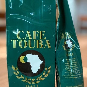 May include: Two green bags of Dali Cafe Touba coffee. The bags feature a white outline of the African continent with a black sun in the upper left corner. The text "CAFE TOUBA" is in gold lettering. The bag on the left has the text "DALI" and "poids net 250 g e" in gold lettering. The bag on the right has the text "DALI" in gold lettering and includes information about the coffee blend, company contact information, and website address.