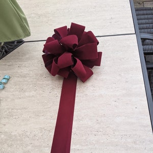 May include: A large, burgundy bow with a long, flowing tail. The bow is made of a soft, velvety material and is arranged in a full, round shape. The tail extends downward, creating a decorative accent. This bow is suitable for gift wrapping or event decorations.