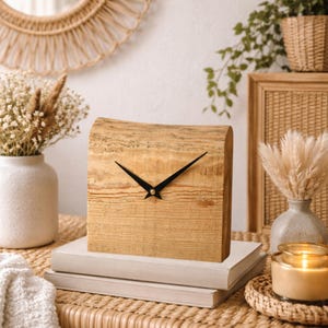 Puede incluir: Un reloj de bloque de madera con manecillas negras se encuentra sobre una pila de libros. El reloj tiene un acabado de veta de madera natural y una forma cuadrada. El fondo incluye un espejo tejido, plantas y objetos decorativos.