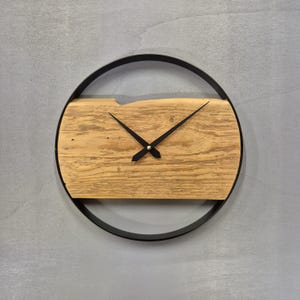Op de afbeelding: Ronde wandklok met een lichtbruine houten wijzerplaat en een zwart metalen frame. De wijzers zijn zwart. De houten wijzerplaat heeft zichtbare nerven. De diameter van de klok is ongeveer 30 cm.