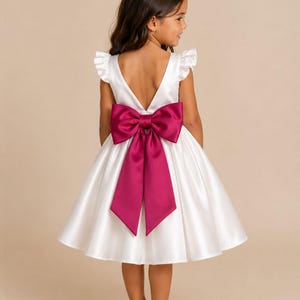 Pode incluir: Um vestido de cetim branco com decote em V profundo nas costas, mangas com babados e saia rodada. Um grande laço magenta vibrante está preso na cintura, com duas fitas longas e largas que caem pelas costas do vestido sobre a saia.