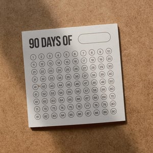 Op de afbeelding: Een vierkant wit papieren notitieblok met de zwarte tekst 90 DAYS OF gevolgd door een leeg rechthoekig vak en een raster van negentig kleine cirkels, elk met een opeenvolgend nummer van één tot negentig in een zwart schreefloos lettertype.