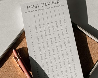 May include: A white paper habit tracker sheet with the text HABIT TRACKER at the top, featuring columns for months and rows numbered 1 through 31 with circular checkboxes, accompanied by a dusty rose colored pen with gold accents.
