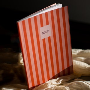 Op de afbeelding: Een notitieboek met een verticaal gestreept patroon in tinten oranje en lichtrood. De voorkant heeft een wit rechthoekig label in het midden met het woord NOTES gedrukt in een donker schreeflettertype. Het notitieboek staat bovenaan open.