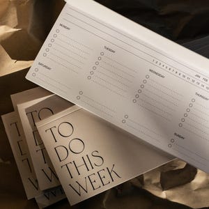 May include: A white weekly planner notepad with black text reading TO DO THIS WEEK on the cover. The interior pages feature a horizontal layout with sections for Monday through Sunday, including dotted lines and circular checkboxes for task tracking.