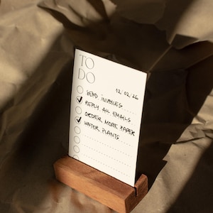 Könnte beinhalten: Eine rechteckige weiße Papierkarte mit einer Aufgabenliste und dem Text TO DO, 12/02/26, SEND INVOICES, REPLY ALL EMAILS, ORDER MORE PAPER und WATER PLANTS, die aufrecht in einem kleinen, rechteckigen braunen Holzblock steht.