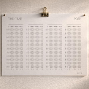 Puede incluir: Calendario minimalista de 2026 impreso en papel crema, sujeto con un clip dorado. El calendario presenta una cuadr&iacute;cula de c&iacute;rculos para cada d&iacute;a, con el texto "THIS YEAR" y "2026" en la parte superior. Montado sobre una superficie texturizada blanquecina.