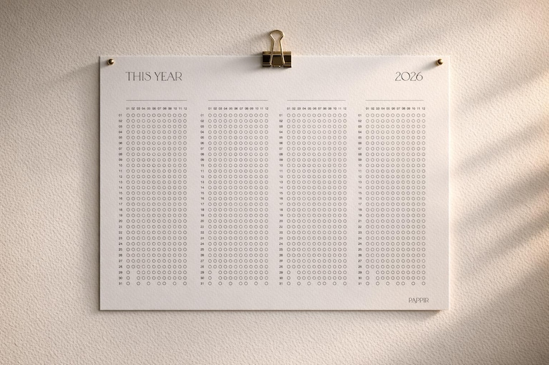 Puede incluir: Calendario minimalista de 2026 impreso en papel crema, sujeto con un clip dorado. El calendario presenta una cuadr&iacute;cula de c&iacute;rculos para cada d&iacute;a, con el texto "THIS YEAR" y "2026" en la parte superior. Montado sobre una superficie texturizada blanquecina.