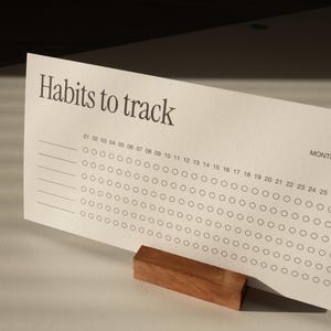 Può includere: Una scheda cartacea color crema per il monitoraggio delle abitudini con il testo "Habits to track" stampato in grigio scuro. La scheda presenta file di cerchi numerati da 01 a 31 per il monitoraggio giornaliero ed è retta da un blocco di legno.