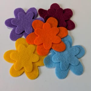 May include: A selection of felt flower cutouts in shades of purple, orange, yellow, blue, and burgundy. The flowers are displayed on a white surface, highlighting their bright colours and simple design. These craft supplies are suitable for various DIY projects.