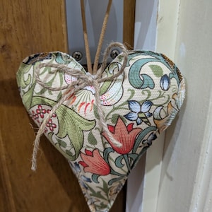 May include: A heart-shaped doorstop made of fabric, featuring a floral design in green, red, and blue on a cream backdrop. It's adorned with twine and a loop for hanging, perfect for a decorative touch.