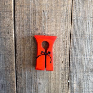 May include: A bright red, wooden ornament shaped like a life vest, with a black string detail. The ornament hangs from a silver wire loop against a weathered wood background. The design is simple and nautical.