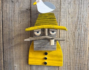 Könnte beinhalten: Eine hölzerne Wandfigur einer Person mit gelbem Hut und gelbem Mantel mit drei schwarzen Knöpfen. Die Figur hat einen Schnurrbart aus Treibholz, eine kleine Pfeife und eine weiß-graue Möwe, die auf dem gelben Hut sitzt.