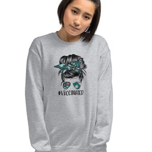 May include: Grey sweatshirt featuring a graphic of a woman with a messy bun and sunglasses. The text "#VACCINATED" is printed below the graphic.