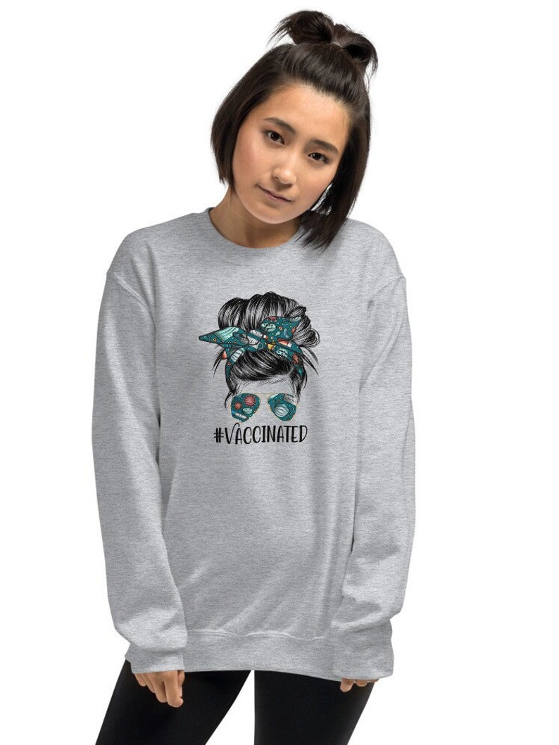 May include: Grey sweatshirt featuring a graphic of a woman with a messy bun and sunglasses. The text "#VACCINATED" is printed below the graphic.