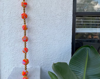 May include: A decorative garland with bright orange pom-poms, accented with purple and yellow, and wooden beads. The garland hangs vertically against a textured white wall. A window and greenery are visible in the background.
