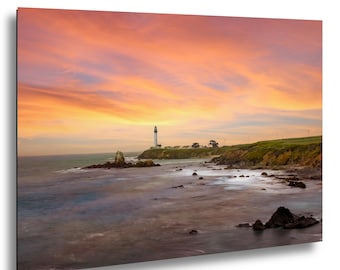 May include: A landscape photograph featuring a tall, slender lighthouse situated on a grassy coastal cliff overlooking the ocean. The scene is illuminated by a vibrant sunset with streaks of orange, pink, and yellow clouds across the sky.