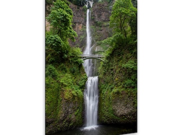 May include: A tall, two-tiered waterfall cascades down a steep, mossy rock cliff face. A white arched stone bridge spans the gap between the two sections of the waterfall, surrounded by lush green trees and dense vegetation on both sides.