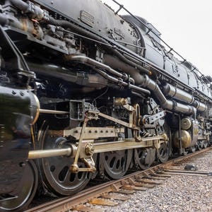 May include: A black steam locomotive with large wheels and intricate piping. The locomotive is on a set of railroad tracks with gravel.