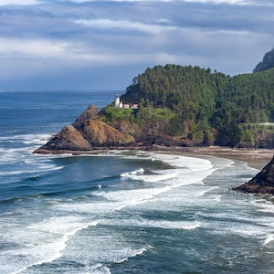 May include: A white lighthouse with a red roof sits on a rocky cliff overlooking a sandy beach and the ocean. The waves are crashing on the shore and there are trees in the background.
