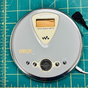 May include: A silver Sony Walkman CD player with a circular design, featuring a central LCD screen, the text Atrac3plus MP3, and the psyc logo. A black power cord is attached. A yellow ruler shows the device is approximately 5.5 inches wide.