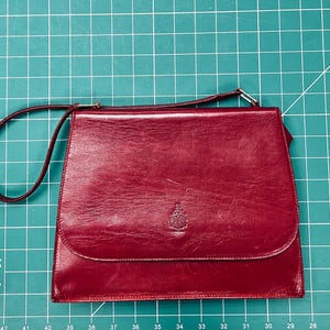 Può includere: Una borsa a tracolla rosso scuro con una finitura liscia e lucida e una forma rettangolare strutturata. La borsa presenta un cinturino sottile, una chiusura a patta e un piccolo logo a stemma in rilievo al centro della patta frontale.