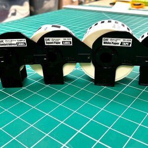 May include: A selection of black label dispensers holding rolls of white paper tape and labels. The labels include text such as "White Paper Tape", "Standard Address Label", and "Square Paper Label". The items are on a green grid surface.