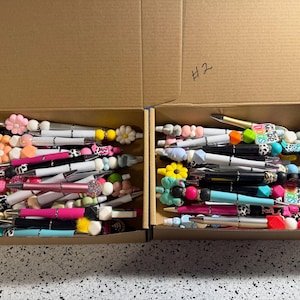 May include: Two open cardboard boxes labeled #1 and #2, each filled with a collection of ballpoint pens. The pens feature various colorful silicone beads, including flower shapes, animal prints, and solid colors, attached to the pen barrels.