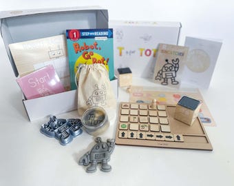 May include: A children's educational toy subscription box with a robot theme. The box includes a book titled "Robot, Go Bot!" a wooden game board, a robot-shaped cookie cutter, and a small bag with a robot design.