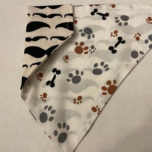 May include: A reversible dog bandana with two different patterns. One side features black moustaches on a cream background. The other side features brown paw prints and black bone shapes on a white background.