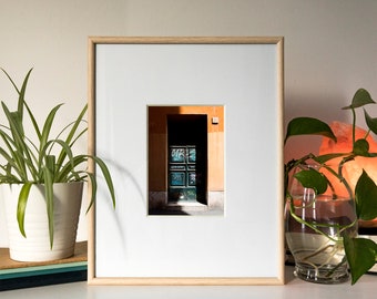 May include: A framed photograph of a doorway with a blue door and graffiti on it. The door is set in a building with orange walls. The photograph is in a light wood frame.