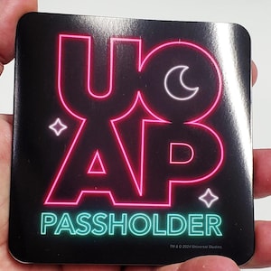 May include: A black square sticker with a pink neon sign that says "YOU ARE A PASSHOLDER" with a crescent moon and two stars. The text "TM & © 2024 Universal Studios." is at the bottom.