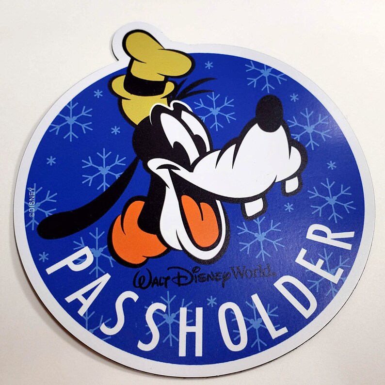 May include: A blue and white round magnet with a cartoon image of Goofy with a red nose and a yellow hat. The text "PASSHOLDER" is written in white letters around the image. The text "Walt Disney World" is written in white letters below the image. The background is blue with white snowflakes.