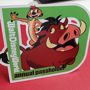 May include: A green and white sticker with a cartoon image of Timon and Pumbaa from Disney's The Lion King. The sticker says "Walt Disney World annual passholder" and includes the Disney FAC number 042894-24183.