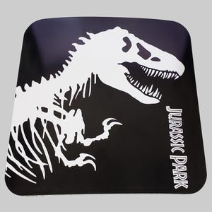 May include: Black square coaster with a white silhouette of a dinosaur skeleton and the text "Jurassic Park" in white.