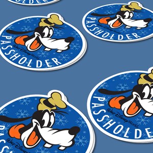 May include: A round blue sticker with a white border features a cartoon image of Goofy wearing a hat and smiling. The background is a blue snowflake pattern. The text "WALT DISNEY WORLD" and "PASSHOLDER" are printed in white on the sticker.