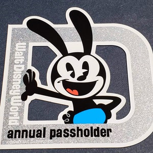 May include: A silver and black sticker with a cartoon character with large ears and a big smile. The character is wearing a blue shirt and the text "annual passholder" is printed below the character. The text "Walt Disney" is printed on the left side of the sticker.