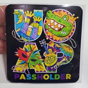 May include: A black square magnet with the letters "U-O-A-P" in colorful, stylized fonts. Each letter is decorated with Mardi Gras-themed imagery, including a crescent moon, a green alligator, a mask, a guitar, maracas, a drum, and a trumpet. The text "PASSHOLDER" is printed in rainbow colors at the bottom.