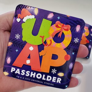 May include: A purple square sticker with a green "U", an orange "A", and a red "P" with a Christmas wreath design. The text "PASSHOLDER" is below the letters and "TM & © 2023 UNIVERSAL STUDIOS." is below the text.