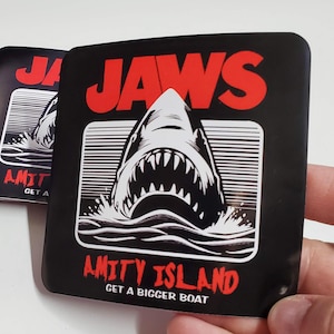 May include: Black square sticker featuring the word "JAWS" in red, above a white and black illustration of a shark's open mouth emerging from the water. Below, the text reads "AMITY ISLAND" and "GET A BIGGER BOAT" in red.